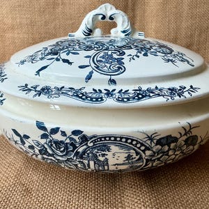 Puede incluir: Una fuente de cer&aacute;mica blanca y azul con tapa. La fuente presenta un dise&ntilde;o floral y paisaj&iacute;stico en azul. Las asas y el adorno de la tapa son ornamentados. La fuente est&aacute; sobre una superficie de arpillera.
