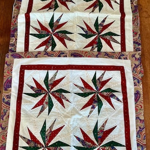 Può includere: Due pezzi di tessuto trapuntato quadrato con un motivo a stella a girandola in rosso, verde e stampa paisley su sfondo bianco. Ogni pezzo è incorniciato da un bordo rosso con piccoli punti bianchi e un bordo esterno in tessuto paisley colorato.