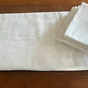 Op de afbeelding: Een wit tafelkleed en een stapel bijpassende servetten met een subtiel, herhalend bloemenpatroon. Zowel het tafelkleed als het bovenste servet zijn geborduurd met de witte monogramletters DW in een decoratieve, cursieve schrijfstijl.