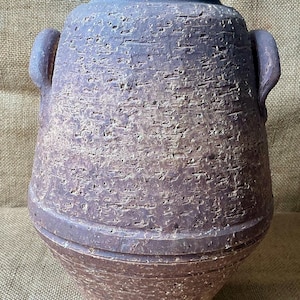 May include: A dark brown, textured ceramic vase with a wide, rounded body, a narrow neck, and two small side handles. The surface features a rough, pitted, and weathered appearance with horizontal ridges near the base and a thick, flat rim.