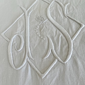 May include: A white linen textile featuring a large, intricate embroidered monogram in white thread. The design consists of stylized, overlapping cursive letters enclosed within a diamond-shaped border, with a small floral sprig detail centered.