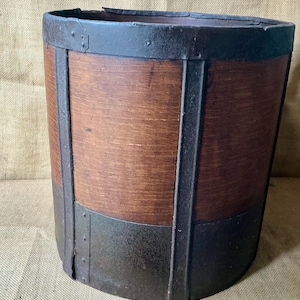 Puede incluir: Un cubo cilíndrico de madera con paneles de madera de color marrón oscuro reforzados con bandas de metal negro en la parte superior, inferior y verticalmente a los lados. Las bandas de metal están aseguradas con pequeños remaches, creando una estructura robusta.