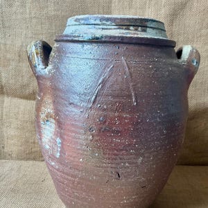 Op de afbeelding: Een grote, verweerde bruine keramische pot met een dikke, afgeronde rand en twee handvatten aan de zijkanten. Het buitenoppervlak heeft een ruwe, getextureerde afwerking met zichtbare horizontale groeven, een vage ingesneden driehoeksmarkering en vlekken van lichtgekleurde minerale afzettingen.