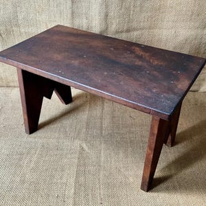 May include: A small, rectangular wooden stool with a dark brown, weathered finish. The stool features a flat, solid top surface and four angled legs that provide stability. The wood grain is visible, showing signs of age and use throughout.