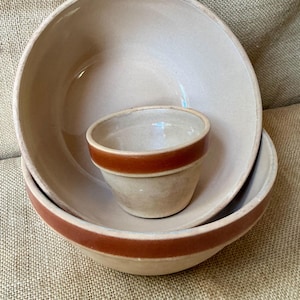 May include: A set of two beige ceramic bowls and one smaller matching ceramic cup. Each piece features a smooth, glossy finish with a distinct, wide horizontal band of reddish-brown glaze encircling the upper exterior rim of the vessels.