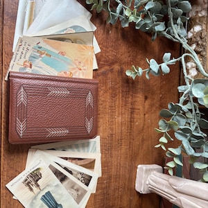May include: A collection of vintage postcards and a brown leather card holder with an arrow design. The postcards feature religious imagery and illustrations. Greenery and a decorative column are also visible, all arranged on a wooden surface.