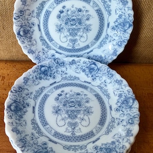 Puede incluir: Una pila de platos de cerámica blanca con un diseño floral azul. Cada plato presenta un ramo central y un borde de rosas y follaje. Los platos tienen un borde festoneado, añadiendo un toque de elegancia a la vajilla.