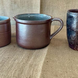Puede incluir: Tres recipientes de cerámica que consisten en dos tazas marrones con asas y una jarra central marrón con asa. Las tazas y la jarra presentan superficies lisas y esmaltadas en varios tonos de marrón, con la taza de la derecha mostrando una textura moteada.