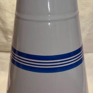 Può includere: Brocca in smalto bianco con bordo blu e tre strisce blu orizzontali. La brocca vintage ha una base leggermente svasata e un beccuccio. Il bordo inferiore mostra segni di ruggine.