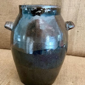 Può includere: Un vaso in ceramica con una smaltatura lucida blu scuro e nera. Il recipiente presenta un corpo largo e arrotondato, un collo corto con un'apertura ampia e due piccole maniglie curve attaccate ai lati superiori. La superficie è liscia e riflettente.