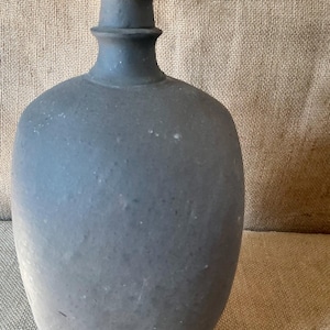 May include: A matte charcoal gray ceramic bottle with a rounded body and a narrow, flanged neck. The surface of the vessel features a textured, slightly mottled finish with subtle variations in tone across the entire exterior of the object.