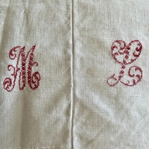 Op de afbeelding: Een witte stoffen ondergrond met twee geborduurde monogrammen in donkerrode draad. De linkerkant toont een gestileerde letter M, terwijl de rechterkant een decoratieve letter L bevat, beide uitgevoerd in een gedetailleerd kruissteekpatroon.