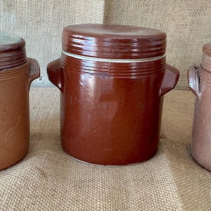 Pode incluir: Três potes cilíndricos de grés com tampas e alças laterais. O pote da esquerda é castanho claro, o central é castanho escuro brilhante e o da direita é castanho médio mate. Cada recipiente apresenta estrias horizontais na parte superior.