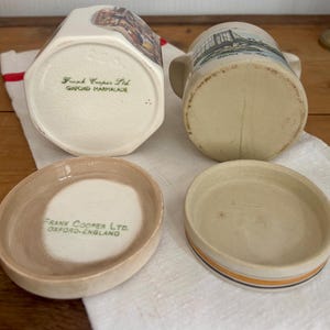 以下が含まれることがあります： 2つのヴィンテージセラミックマーマレードジャーと2つの蓋。1つのジャーは六角形でカラフルなイラストが描かれています。もう1つのジャーは円筒形で橋のイラストが描かれています。蓋は丸く、茶色と白の配色です。「Frank Cooper Ltd. Oxford Marmalade」の文字が見えます。