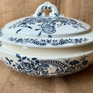Puede incluir: Una fuente de cer&aacute;mica blanca y azul con tapa. La fuente presenta un dise&ntilde;o floral y paisaj&iacute;stico en azul. Las asas y el adorno de la tapa son ornamentados. La fuente est&aacute; sobre una superficie beige.