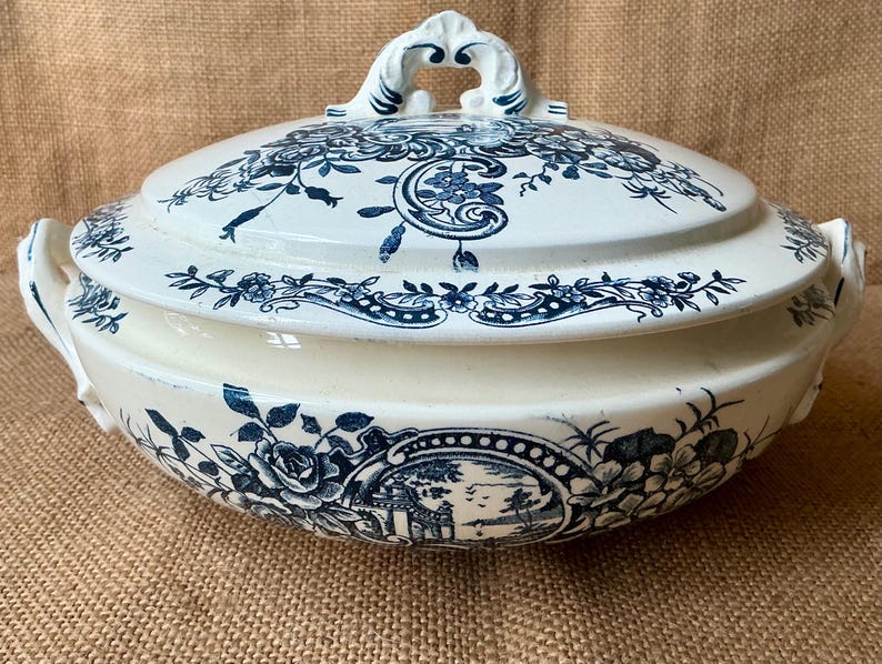 Puede incluir: Una fuente de cer&aacute;mica blanca y azul con tapa. La fuente presenta un dise&ntilde;o floral y paisaj&iacute;stico en azul. Las asas y el adorno de la tapa son ornamentados. La fuente est&aacute; sobre una superficie beige.