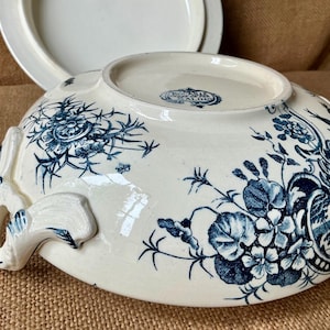 Puede incluir: Cuenco de cer&aacute;mica color crema con motivos florales azules y asas. El cuenco tiene una tapa en el fondo. El dise&ntilde;o floral incluye flores y hojas. El cuenco es probablemente vintage o antiguo.