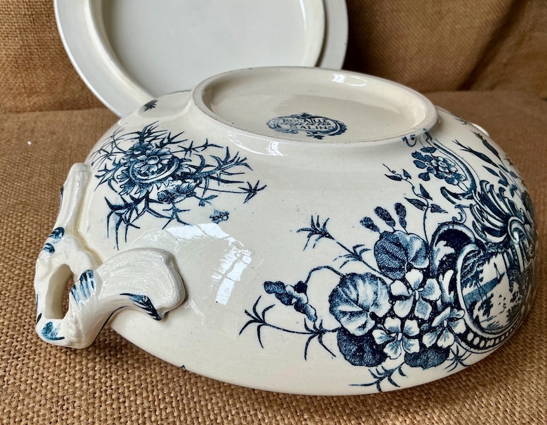 Puede incluir: Cuenco de cer&aacute;mica color crema con motivos florales azules y asas. El cuenco tiene una tapa en el fondo. El dise&ntilde;o floral incluye flores y hojas. El cuenco es probablemente vintage o antiguo.
