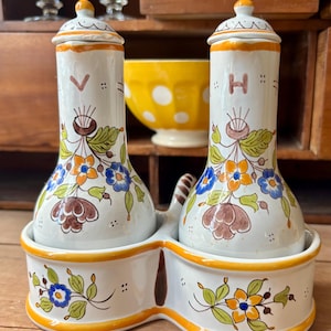 Può includere: Un set di sale e pepe in ceramica bianca con disegni floreali blu, arancioni e marroni. Il set comprende due shaker cilindrici con coperchi, seduti in una base collegata con un bordo giallo. Gli shaker hanno le lettere "V" e "H".