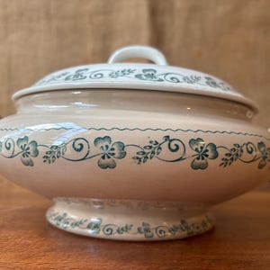 May include: A beige ceramic tureen with a lid and handles. The tureen is decorated with a green floral pattern around the body and lid. The lid has a white handle.