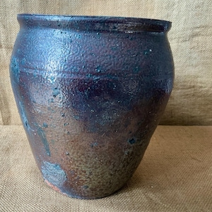 May include: A rounded ceramic pot featuring a textured, mottled glaze in deep shades of dark blue, purple, and earthy brown. The surface has a rough, pitted appearance with subtle variations in colour density and a wide, open circular rim.