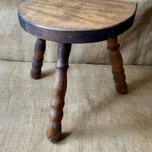 May include: A small, three-legged wooden stool featuring a semi-circular seat with a dark brown finish. The stool is supported by three turned, spindle-style legs that are attached to the underside of the flat, curved wooden seat surface.