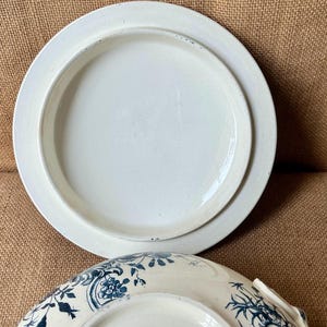 Puede incluir: Dos platos de cer&aacute;mica blanca, uno apilado sobre el otro. El plato superior es liso, mientras que el plato inferior presenta un dise&ntilde;o floral azul. Los platos est&aacute;n sobre una superficie marr&oacute;n.