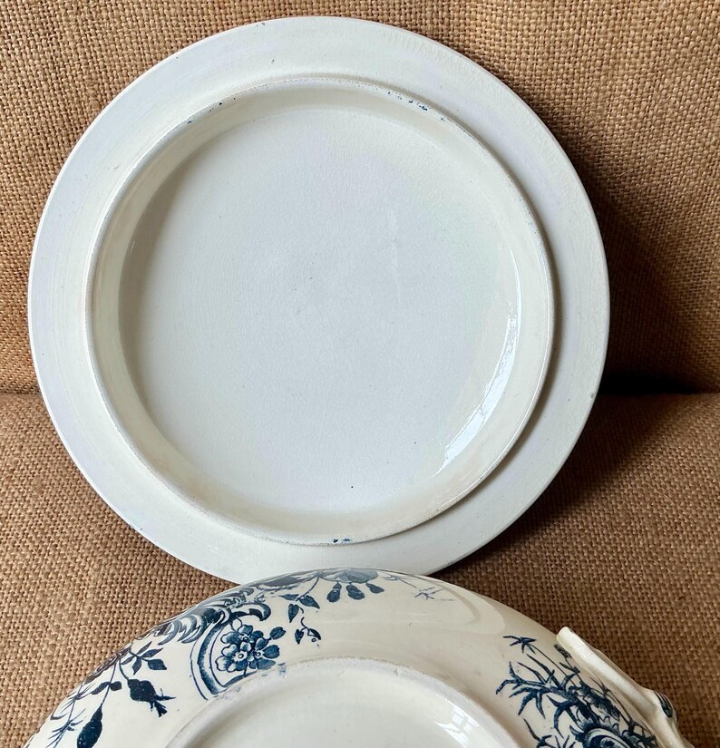 Puede incluir: Dos platos de cer&aacute;mica blanca, uno apilado sobre el otro. El plato superior es liso, mientras que el plato inferior presenta un dise&ntilde;o floral azul. Los platos est&aacute;n sobre una superficie marr&oacute;n.