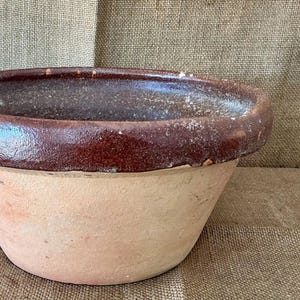 Puede incluir: Un cuenco redondo de barro con un borde ancho y grueso esmaltado en un acabado marrón oscuro brillante. La parte inferior del cuenco permanece sin esmaltar, mostrando una textura de arcilla de color tostado claro mate y natural.