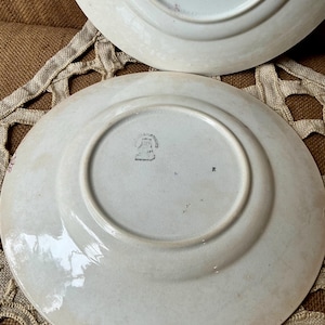 Puede incluir: Dos platos antiguos de cerámica blanca con un tono ligeramente blanquecino. Los platos tienen un diseño simple y clásico con un borde elevado y un área central empotrada. La parte inferior de un plato muestra un sello descolorido y la letra "R".