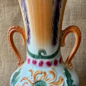 Può includere: Un vaso decorativo in ceramica con la parte superiore svasata e due manici curvi. Il vaso presenta un motivo floreale con accenti arancioni, verdi e rosa. La combinazione di colori comprende arancione, viola e bianco.