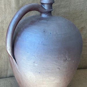 Op de afbeelding: Een grote, ronde keramische kan met een donkerbruine, gevlekte afwerking. De kan heeft één gebogen handvat bevestigd aan de hals en schouder, een smalle tuit met een uitlopende rand en subtiele horizontale ribbels rond het bovenlichaam.