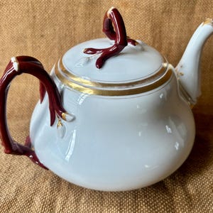 May include: A white porcelain teapot with a rounded body, a curved spout, and a lid. The handle and lid finial are a dark burgundy colour. The rim and accents are trimmed with gold. A classic tea accessory.