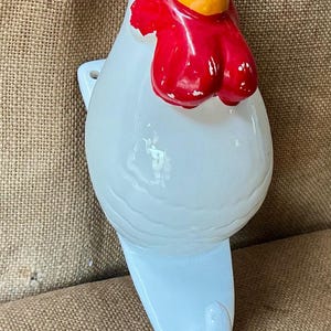 Puede incluir: Un gancho de pared de cerámica blanca con forma de gallo. El gallo tiene una cresta y barbillas de color rojo brillante, un pico naranja y un ojo negro. El cuerpo es liso y blanco, con un gancho curvado en la parte inferior para colgar objetos.