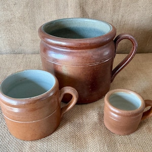 Könnte beinhalten: Ein Set aus drei braunen Keramikgefäßen mit hellblauer Innenseite. Die Sammlung umfasst einen großen Topf mit Henkel, einen mittelgroßen Becher mit Henkel und eine kleine Tasse mit Henkel, alle mit glatter Glasur und zylindrischer Form.