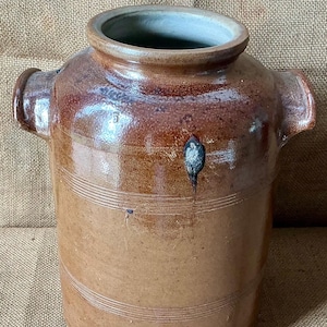 May include: A cylindrical brown stoneware crock featuring a wide mouth, two side handles, and a lower spigot opening plugged with a cork. The exterior has a glossy glaze with horizontal incised lines and a dark spot near the upper rim.