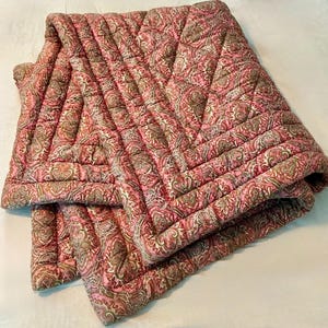 Pode incluir: Uma manta acolchoada com um padrão paisley repetitivo em tons de rosa, verde suave e creme. O tecido é costurado com uma grade de linhas retas, criando uma aparência texturizada e acolchoada em toda a superfície do têxtil.
