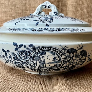 Puede incluir: Fuente de cer&aacute;mica color crema con tapa, adornada con motivos florales y paisaj&iacute;sticos azules. La fuente tiene un asa en la tapa y asas laterales. El dise&ntilde;o incluye rosas y un paisaje.