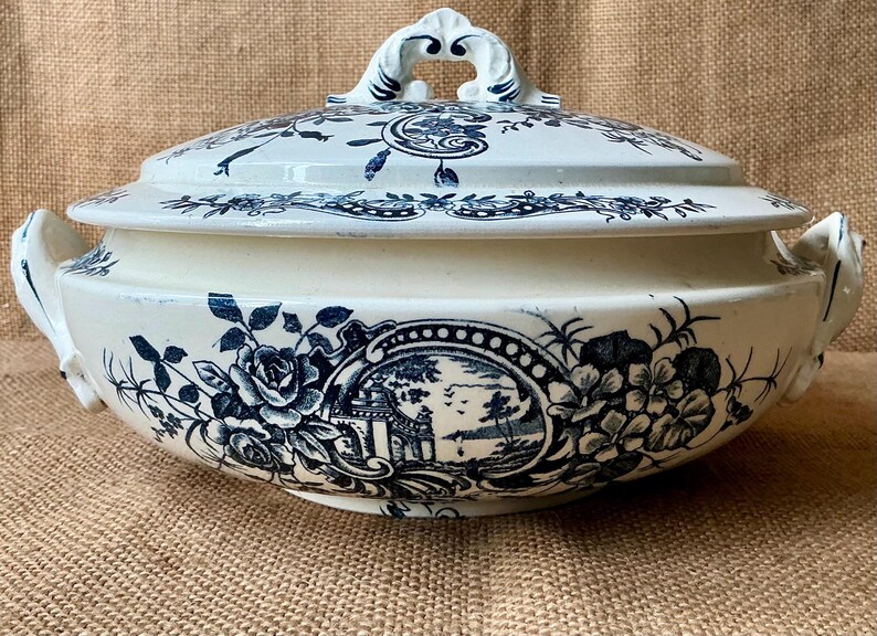 Puede incluir: Fuente de cer&aacute;mica color crema con tapa, adornada con motivos florales y paisaj&iacute;sticos azules. La fuente tiene un asa en la tapa y asas laterales. El dise&ntilde;o incluye rosas y un paisaje.