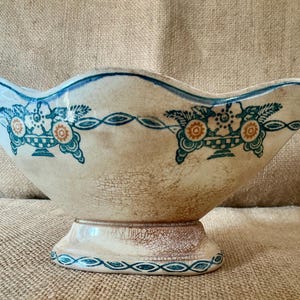 May include: A ceramic bowl with a scalloped rim and a pedestal base. The off-white surface features a visible crackle glaze pattern and a repeating teal and orange floral motif in a basket design, with a teal line accenting the scalloped edge.