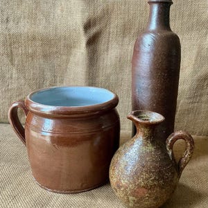 Puede incluir: Una vasija de cerámica marrón con interior azul claro, una botella alta de color marrón oscuro con acabado liso y una pequeña jarra redonda de cerámica moteada en marrón y verde con asa, dispuestas como un conjunto de tres objetos.