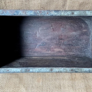 May include: A rectangular wooden brick mold featuring dark brown wood grain and reinforced with weathered, oxidized metal bands along the edges. The metal frame is secured with visible rivets, creating a sturdy, open-top box structure.