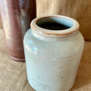 Może przedstawiać: Jasnoszary ceramiczny garnek z szerokim otworem i brązową krawędzią. Z przodu znajduje się wytłoczony kwadrat z napisem H.V PARIS. Wysokie, ciemnobrązowe cylindryczne naczynie ceramiczne jest częściowo widoczne za szarym garnkiem.