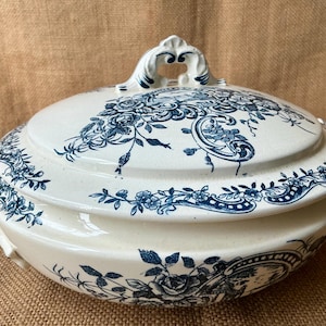 Puede incluir: Una fuente de cer&aacute;mica blanca con tapa, adornada con patrones florales azules. La fuente tiene un asa en la tapa y asas laterales. El dise&ntilde;o floral incluye rosas y otros elementos bot&aacute;nicos, creando una est&eacute;tica cl&aacute;sica y elegante.