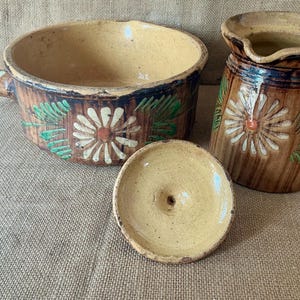 May include: A brown glazed ceramic cooking pot with a handle, a matching lid, and a tall pitcher. Both pieces feature a hand-painted white daisy design with green leaves against a brown, vertically streaked background and a cream-colored interior.