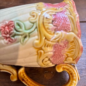 May include: Cream ceramic teacup with embossed floral and scroll designs. The cup features pink flowers, green leaves, and gold accents. The curved handle adds to the vintage aesthetic. A decorative piece.