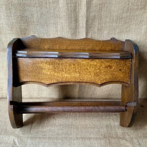 Puede incluir: Un soporte de madera para papel de cocina montado en la pared con un acabado marrón medio y veta de madera visible. La unidad incluye un estante superior plano con un borde festoneado y una varilla horizontal en la parte inferior.