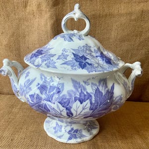 May include: A white ceramic tureen with a blue ivy leaf pattern, featuring two side handles and a matching lid with a decorative loop handle. The vessel has a rounded body and sits on a pedestal base, decorated with blue botanical illustrations.