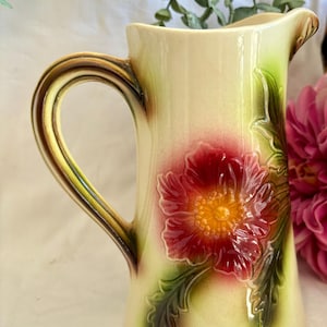 Può includere: Un'antica brocca in ceramica con un motivo floreale. La brocca è di colore crema con un grande fiore rosso con centro giallo e foglie verdi.