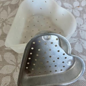 May include: A heart-shaped kitchen tool set, featuring a white plastic container with small holes and a silver metal mold with perforations. The set is designed for food preparation, likely for shaping or steaming.